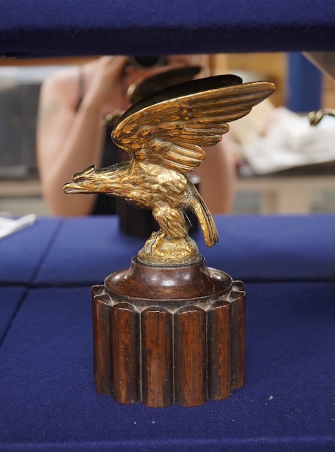 Two brass car mascots in the form of eagles, one mounted on a threaded radiator cap, the other mounted on a wooden base, the larger with a wingspan of 31cm. Condition - fair, one eagle with bent wing.